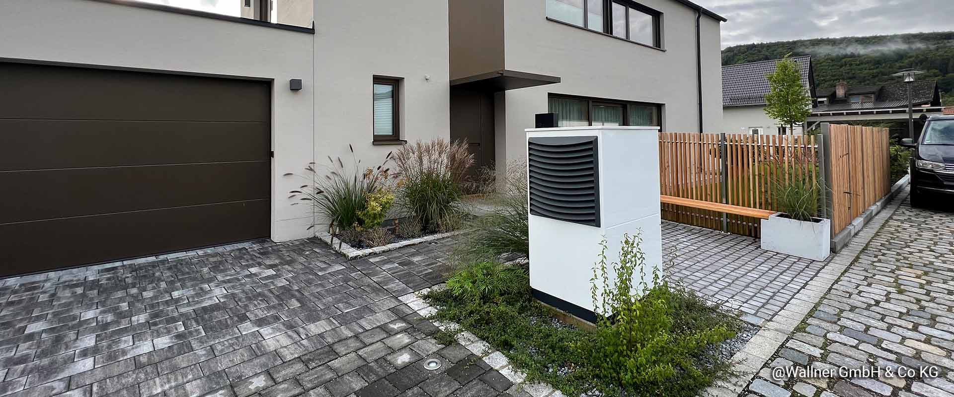 An air source heat pump can be seen installed on some decorative gravel and surrounded by some small plants. A paved way around the heat pump leads to the new building in the background. On the right, there is a fence with some meadow behind.