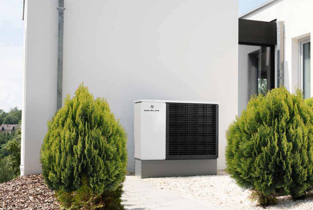 The air/water heat pump LADV is installed outdoors on white decorative stones in front of a house wall, next to a paved way and two small bushes.