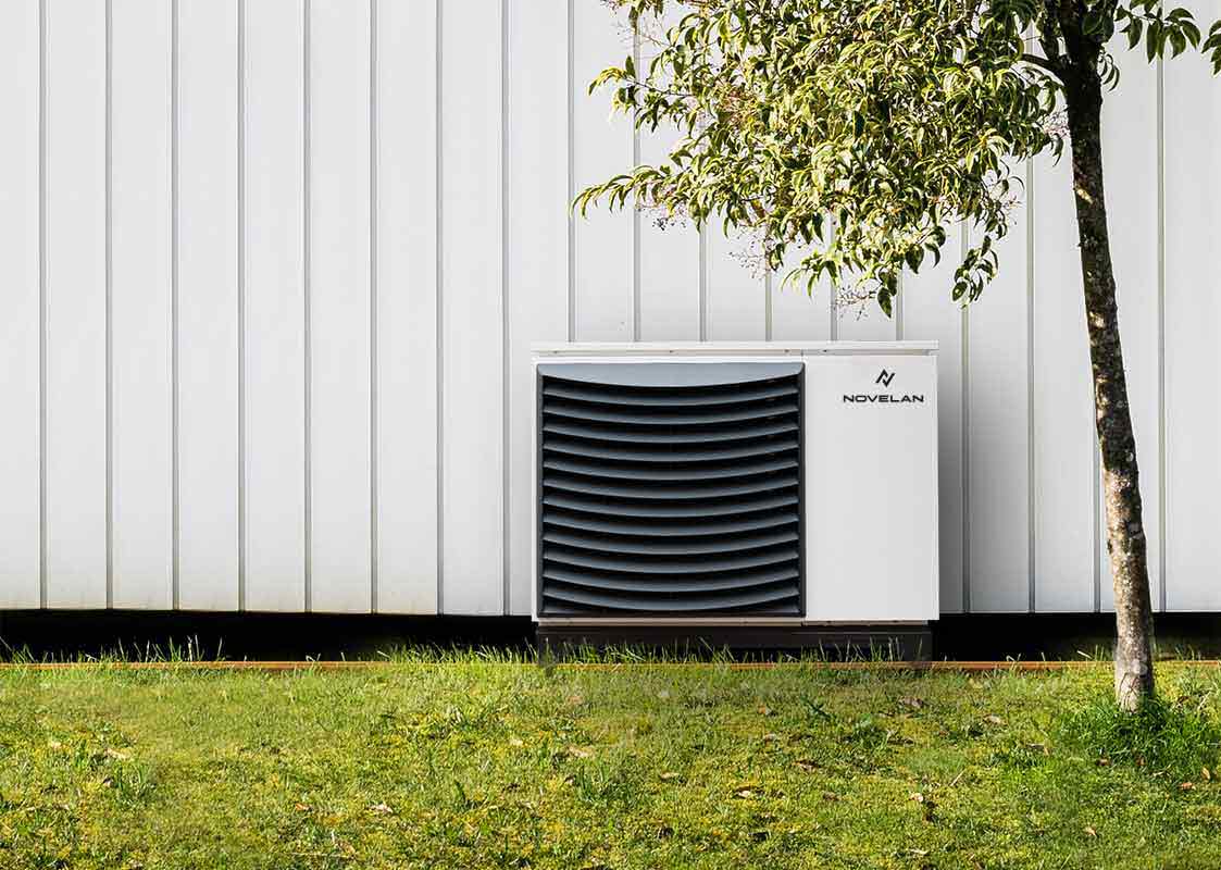 The air source heat pump Jabbah can be seen with a ventilation grille on the left front and the NOVELAN logo in the upper right corner as it stands in front of a wooden house wall next to a tree with a meadow in the foreground.