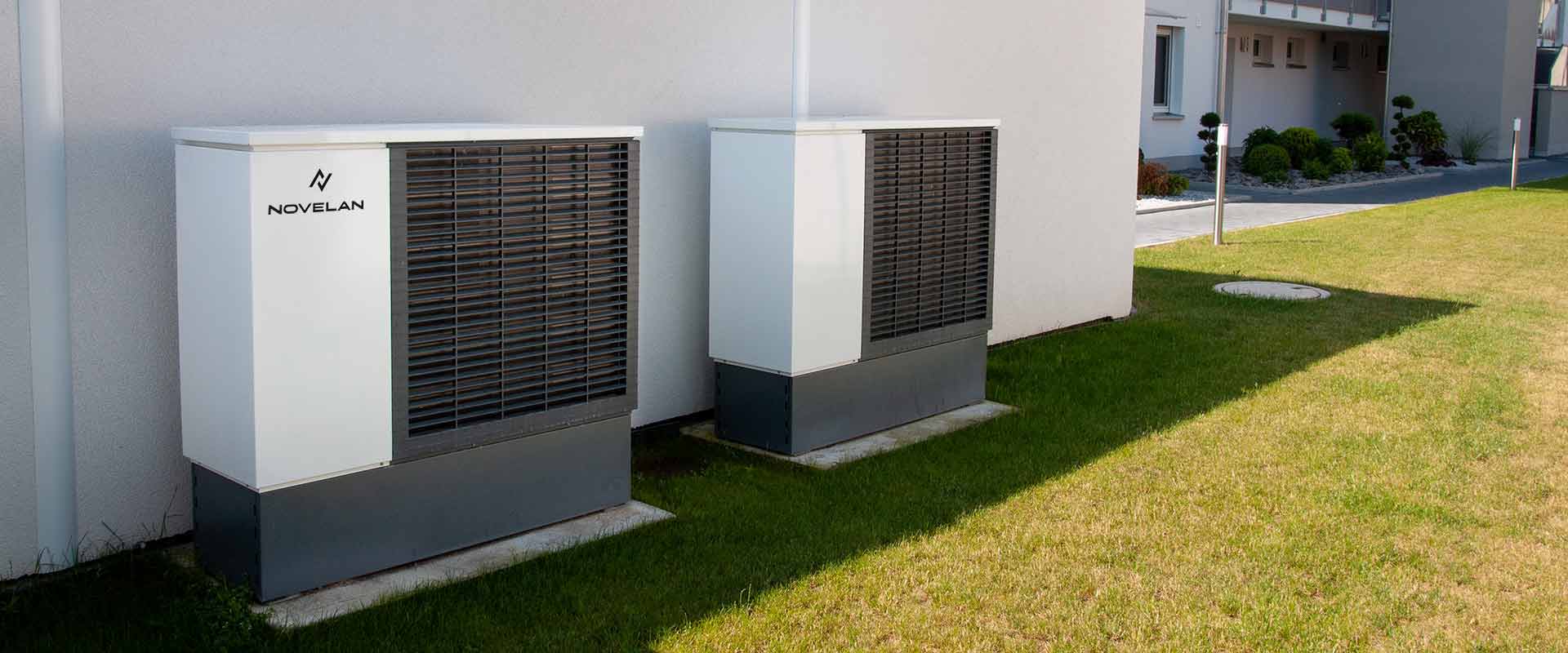 Two LADV heat pumps are installed next to each other in front of a house wall on a meadow. In the background on the right, the entrance and a paved way to the front door can be seen.