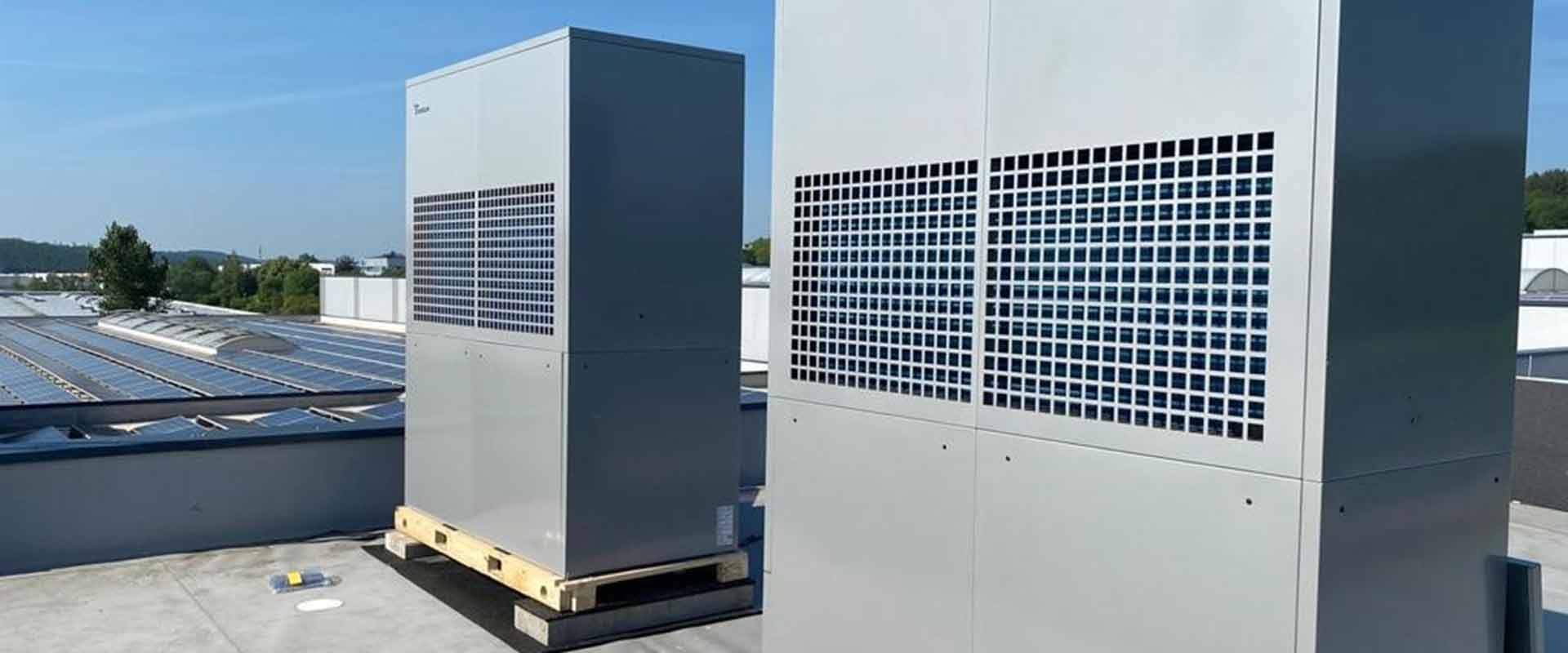 Two LAP heat pumps were photographed diagonally from the right, on the top of a flat factory roof with photovoltaic panels. In the background, blue and cloudless sky can be seen.