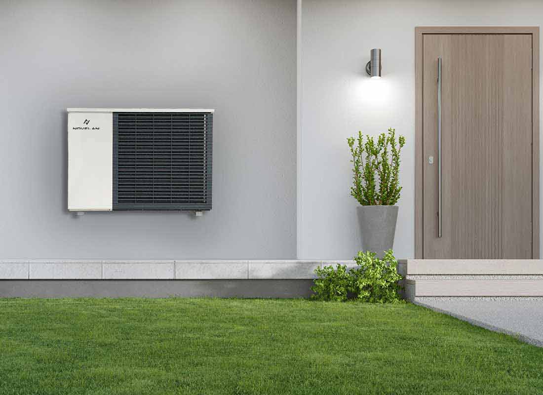 The air source heat pump LAD is installed outdoors on a house wall. On its right, there is the front door which has a potted plant standing on its left and a light on the wall above. In the foreground, there is a meadow and in front of the door, there are stairs with a plant on the left.