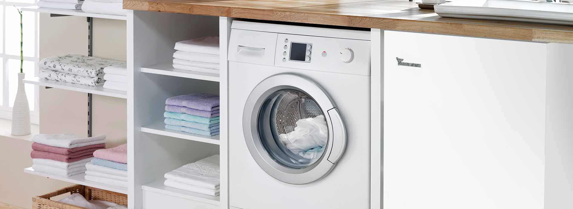 A brine/water-heat pump is installed underneath a work surface in a kitchen next to a washing machine and open cupboards with folded towels on the left.