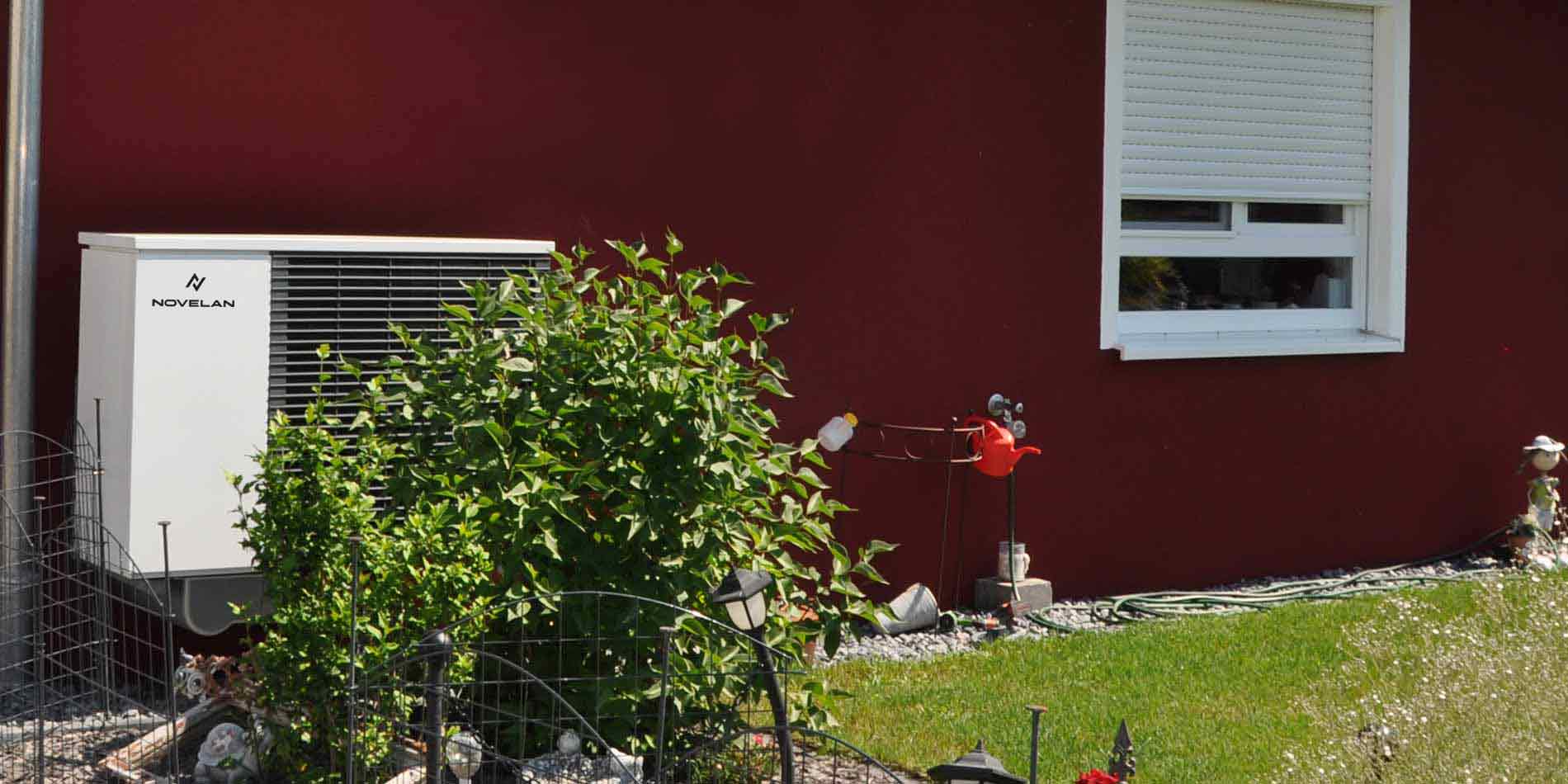 The air source heat pump LADV can be seen outdoors on a house wall above decorative gravel. In the foreground, there is some meadow and a bush on the left covering half of the heat pump.