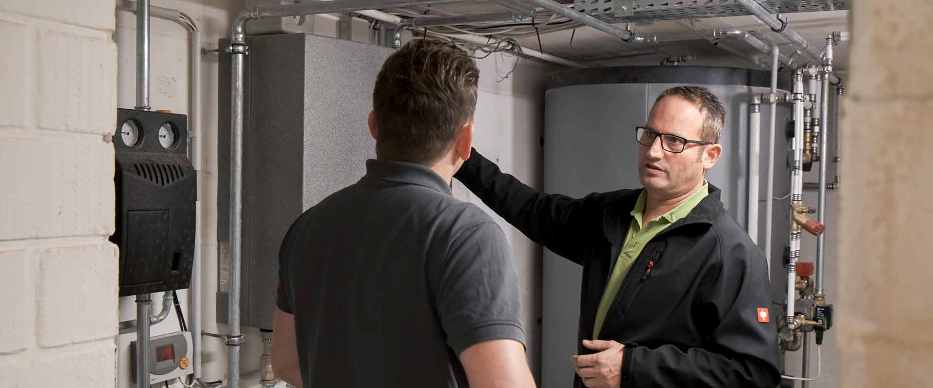 Two men stand in a basement in front of a buffer tank and next to a Hydraulic module as the one in the back, wearing a green polo shirt underneath a black jacket, explains the device to the other man standing in front of him.