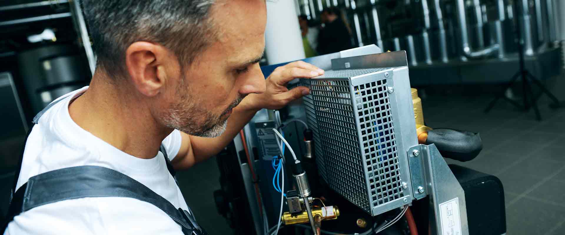 A man can be seen in close-up while working on component parts of a heat pump. Pipes can be seen blurry in the background, extending into the wall.