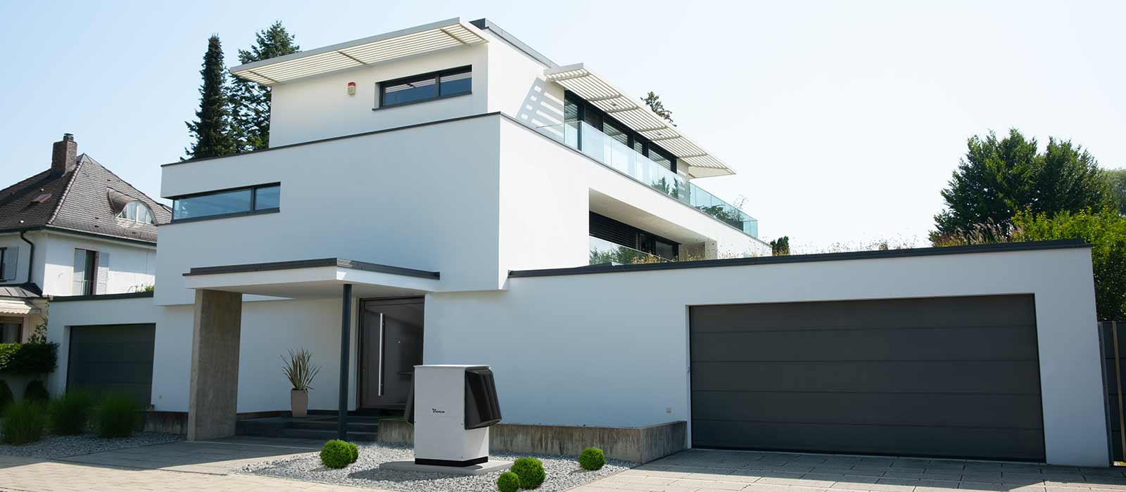 The heat pump LAVS with large sound insulation hoods on both sides stands on decorative gravel with some small bushes in front of a modern residential building with 3 floors. The house got flat roofs, the front door in the middle and two garages on the left and right, respectively.