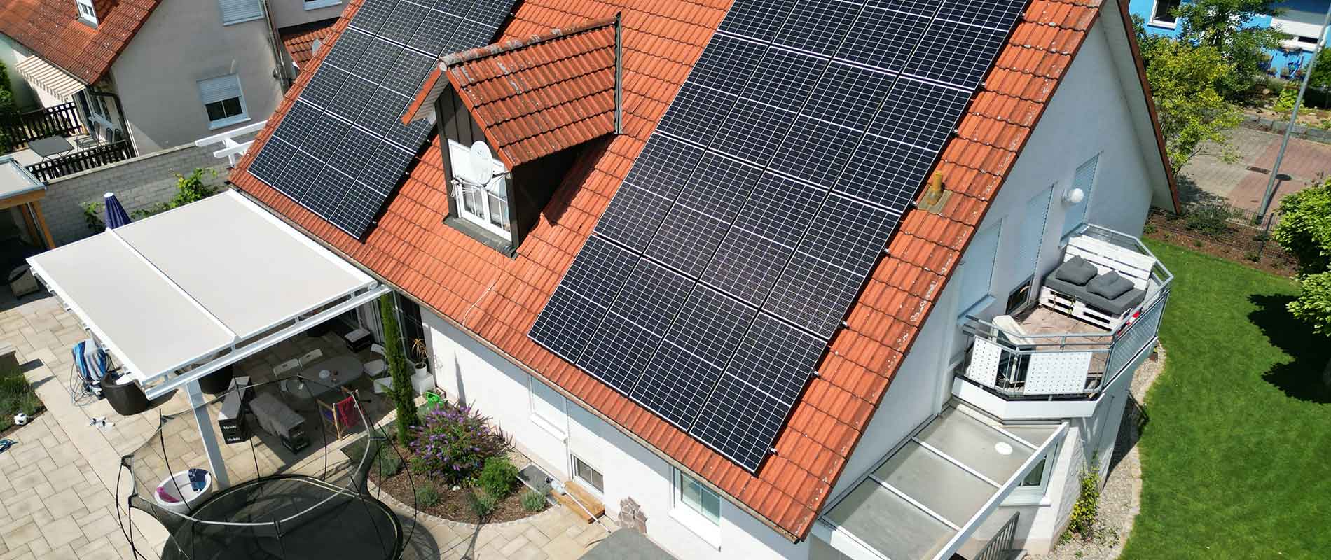 A house with photovoltaics on its sloping roof can be seen from an aerial view, with a terrace on the left side and a balcony on the right side above some green area.