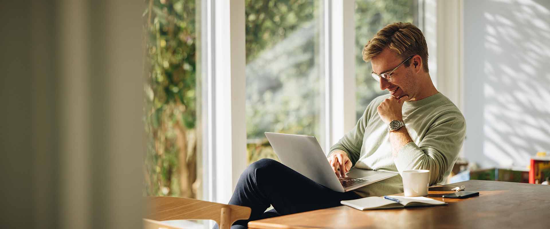 A smiling man sits at a table, on it a mug, open notebook, cell phone and ear pods, while he is watching something on a laptop on his lap. The sun shines through the floor-length windows.