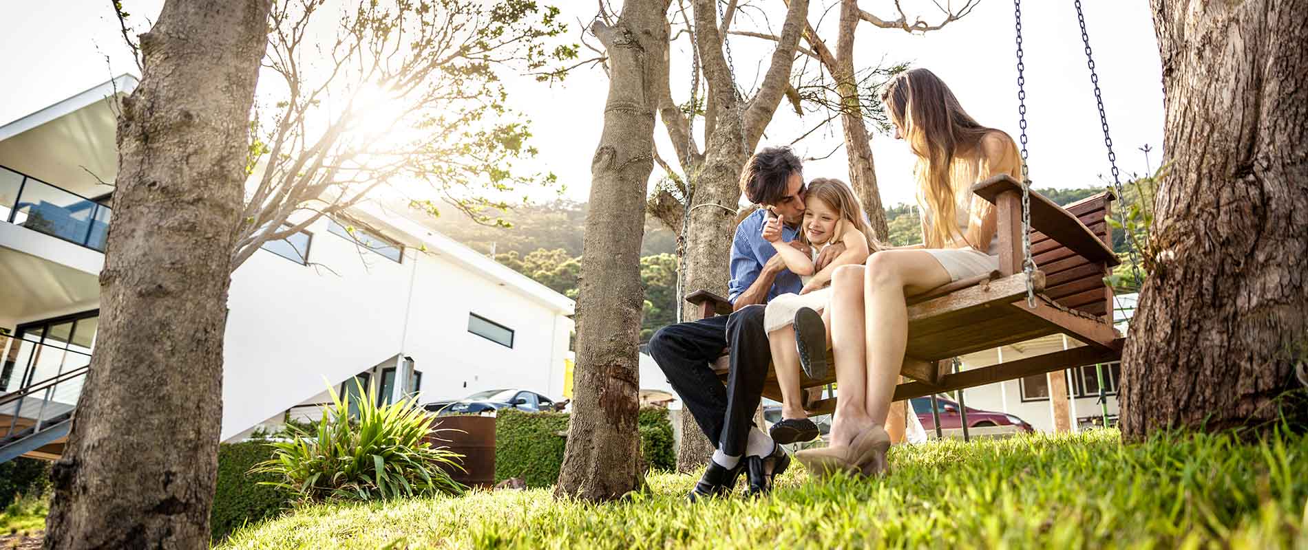 A young family with the child in the middle sits on a Hollywood swing as the father pulls his daughter to the left to kiss her hair while the mother smiles happily. The sun shines through the surrounding trees and a modern apartment building is recognizable in the background with green hills farther behind.