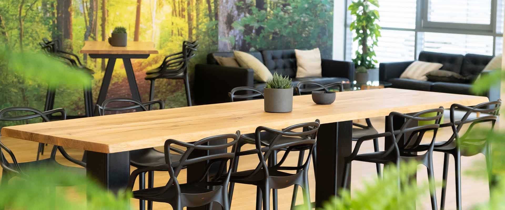 The foyer of the ait-Academy can be seen with a big wooden table and several black chairs. In the background there is another table and two black leather couches in the corner in front of a forest wallpaper. Different plants are put next to the couches and on the tables.