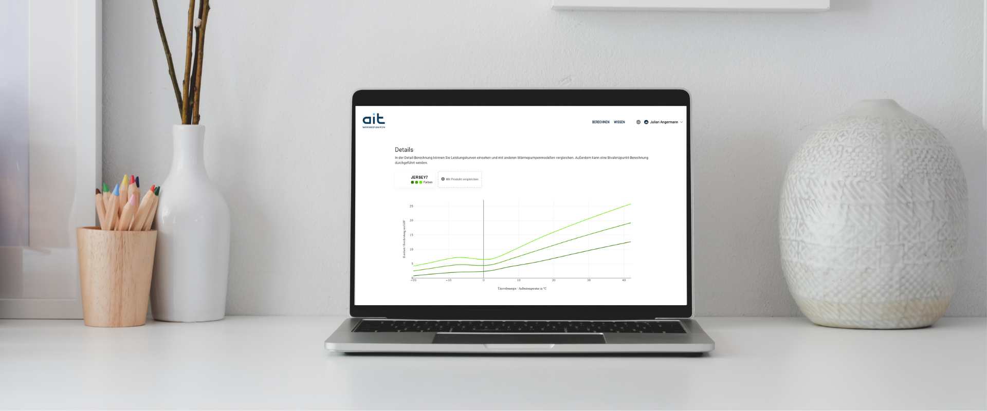 An opened laptop on a table shows a curve diagram on the myait-website about the performance of the heat pump Jersey. On its left there is a cup with colored pencils and a vase on the right side.